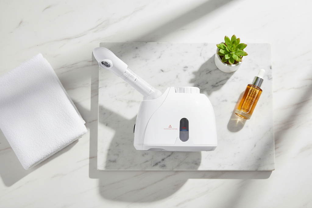 Veluné Aura Deep Clean Facial Steamer flat lay with towel, plant and serum on marble
